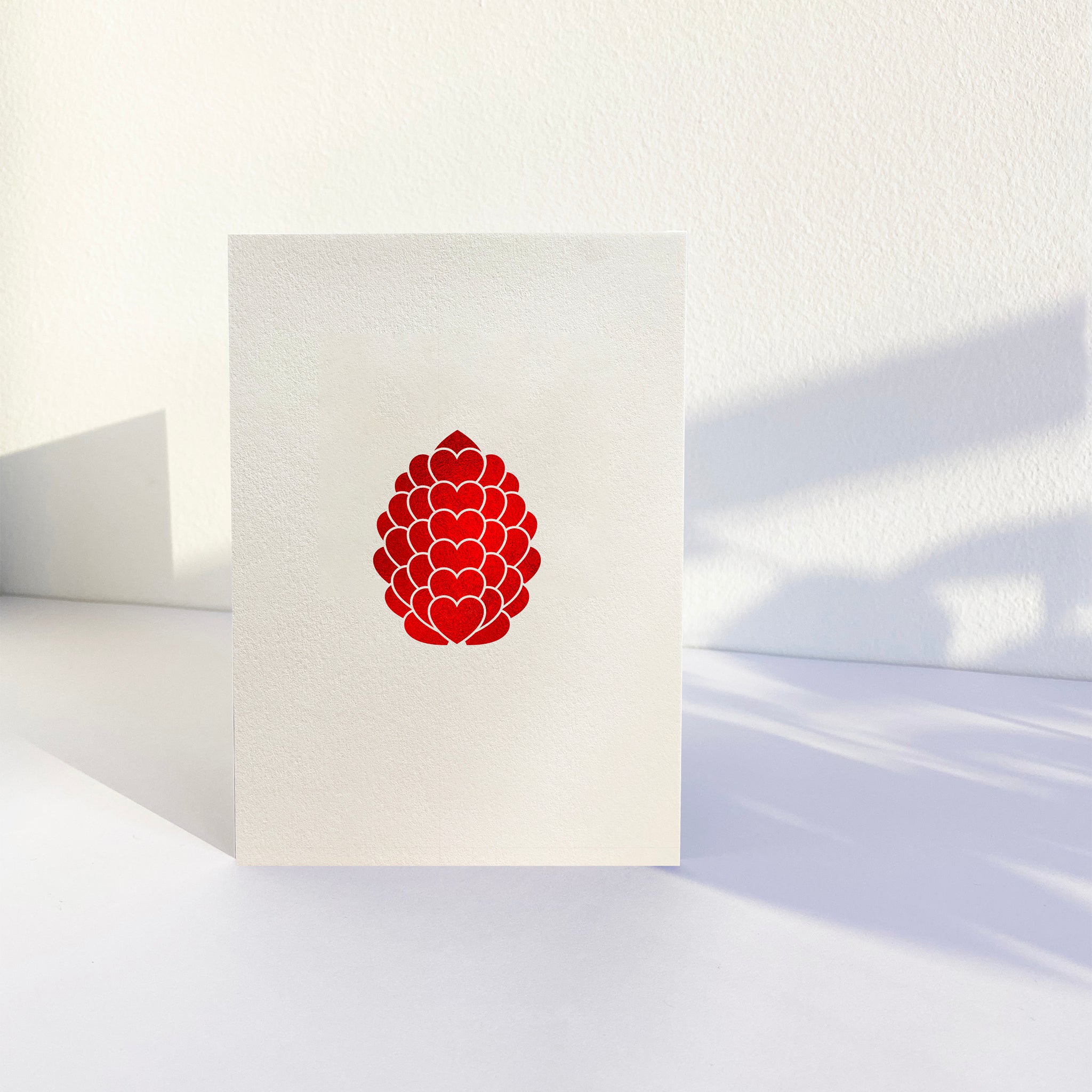 Hot foil pressed pinecone love heart graphic in red on white card. Card is vertical and standing with white background.