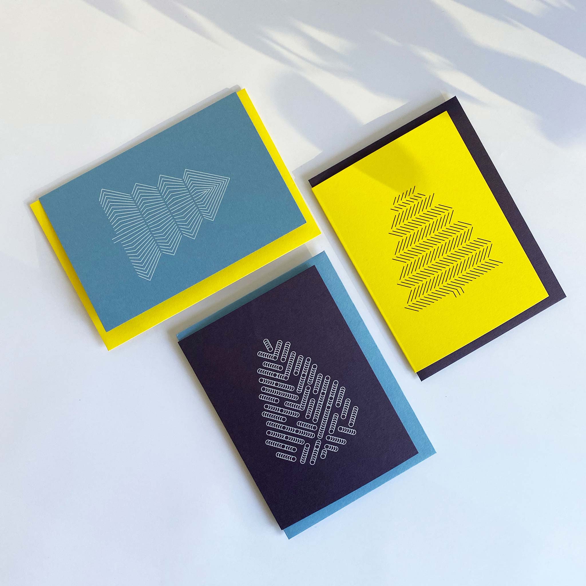 Flat lay of three christmas greeting cards with geometric tree prints. Deep purple with white tree, sky blue with white tree and bright yellow with black tree. Alternating coloured envelopes. 