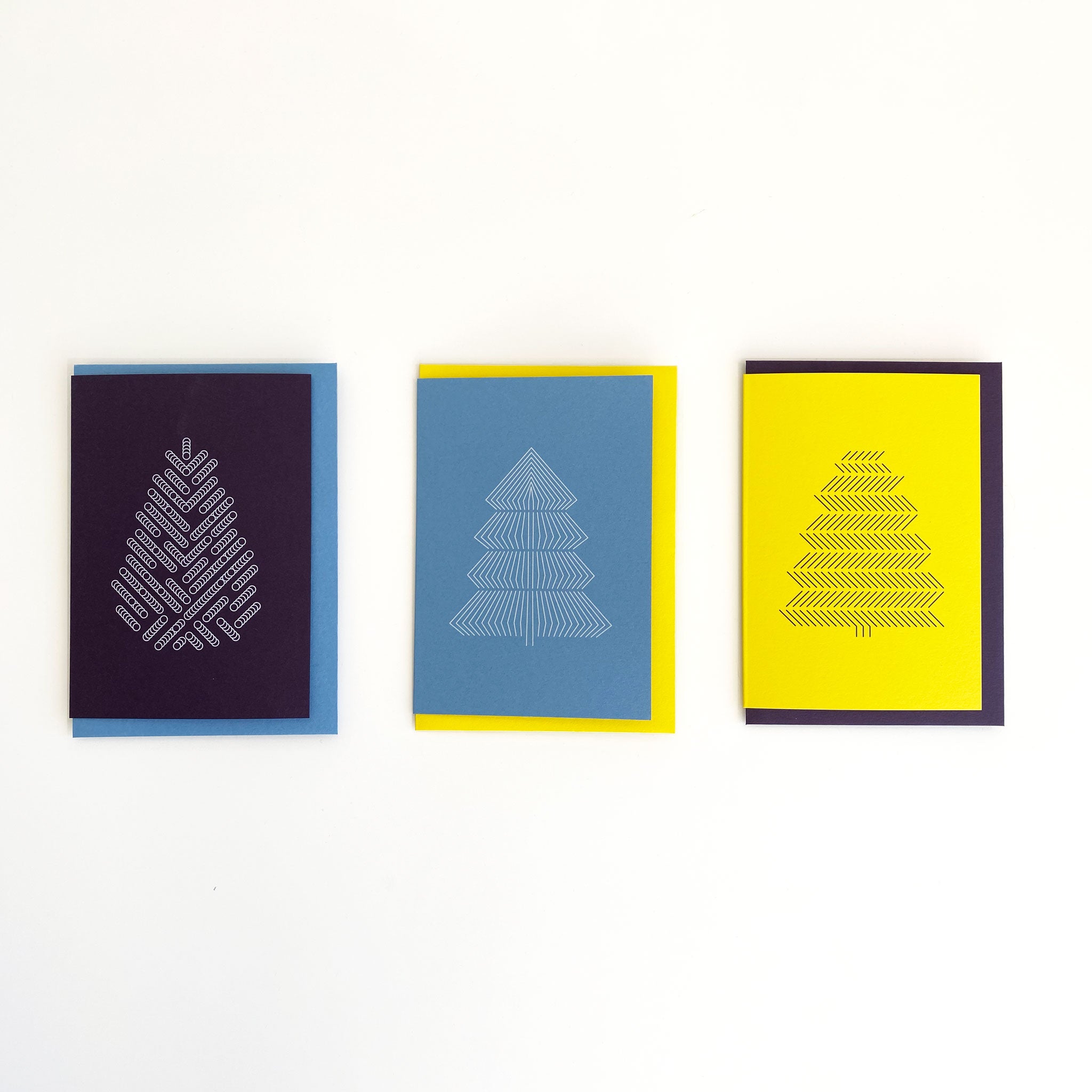 Flat lay of three christmas greeting cards with geometric tree prints. Deep purple with white tree, sky blue with white tree and bright yellow with black tree. Alternating coloured envelopes.