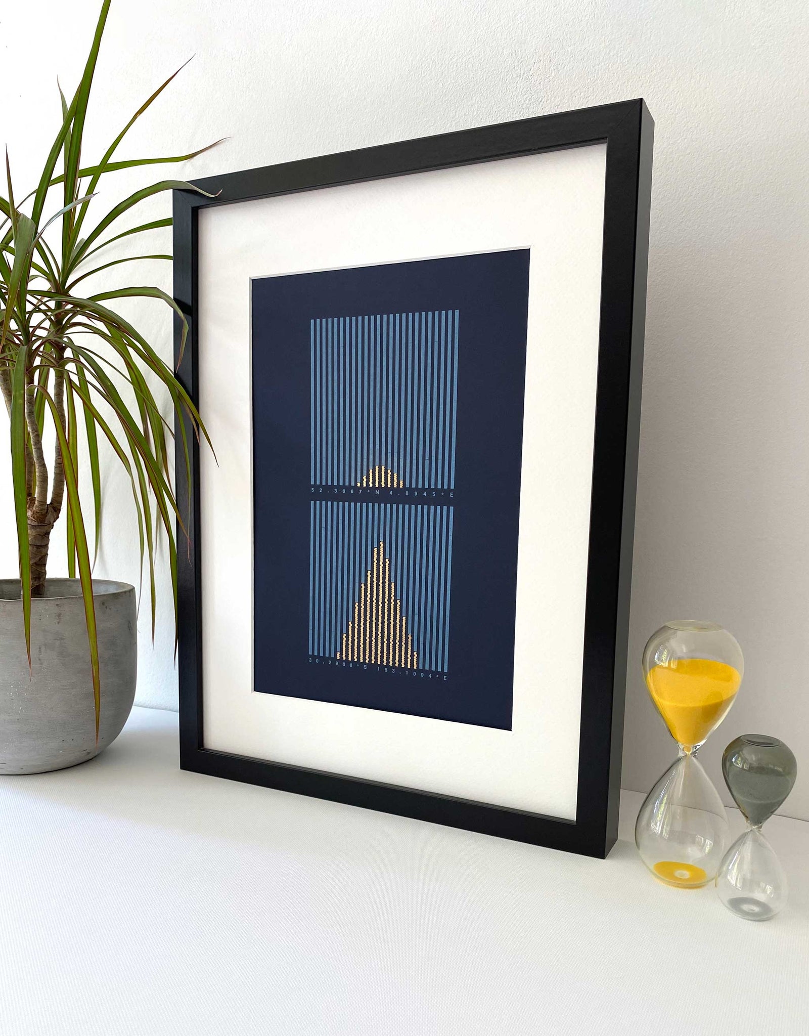 Framed Letterpress data print Sunlight Hours. Light blue and gold vertical lines on dark blue background. Lifestyle image with plant and props.